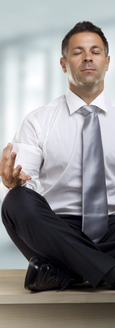 Ein meditierender Mann in mittlerem Alter in Business-Outfit: im Yogasitz, rechte Hand auf Knie abgestützt, Augen geschlossen, in schwarzer Anzughose, weißem Hemd und glänzender silbergrauer Krawatte.