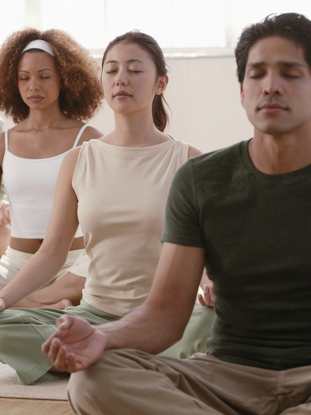 Drei junge Meditierende in Yogasitz am Boden eines Raumes. von vorne rechts nach links hinten: ein indischer Mann, eine japanische Frau, eine Afroamerikanerin.