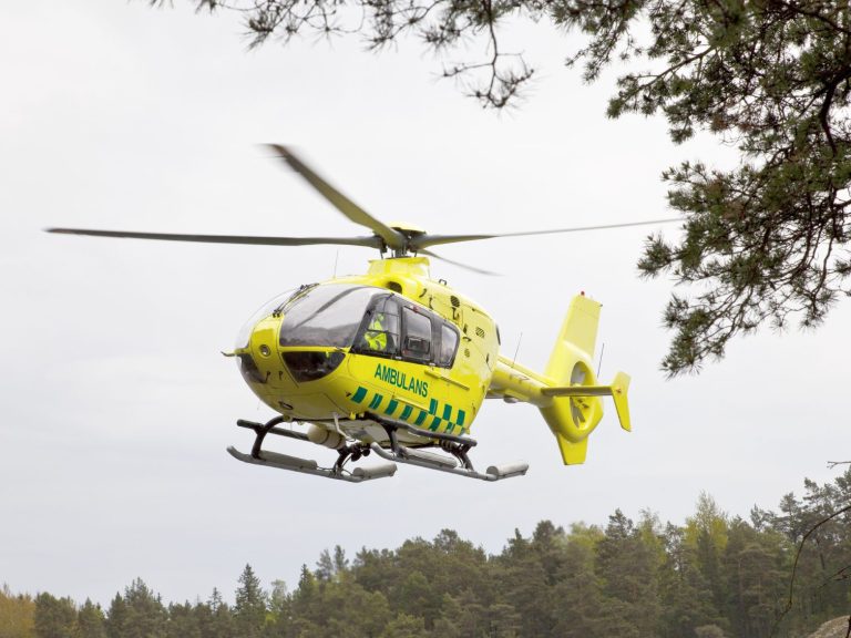 Zitronen-gelber Rettungshubschrauber im Landeanflug auf eine Waldlichtung in hügeligem Gelände.