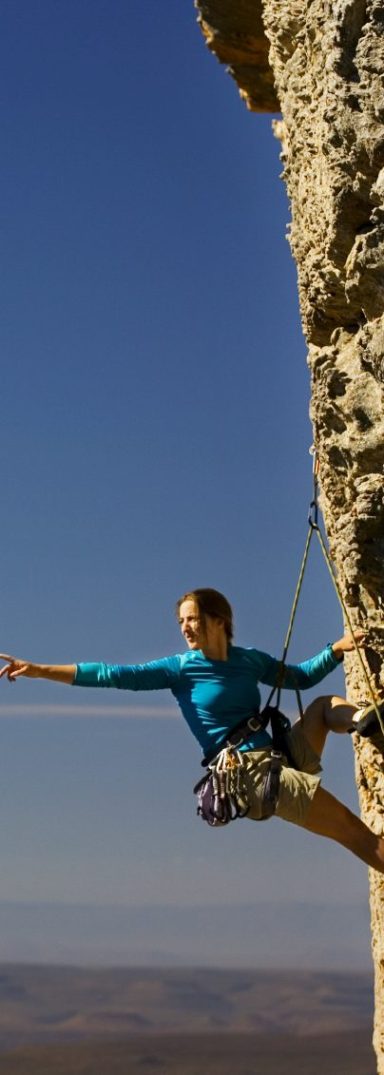 Junge Frau hängt hoch in einer senkrechten Felswand, im Ausfallschritt, den Blick von der Wand weg weit auf den Horizont gerichtet, auf diesen zeigend, gesichert durch Klettergurt und Haken.