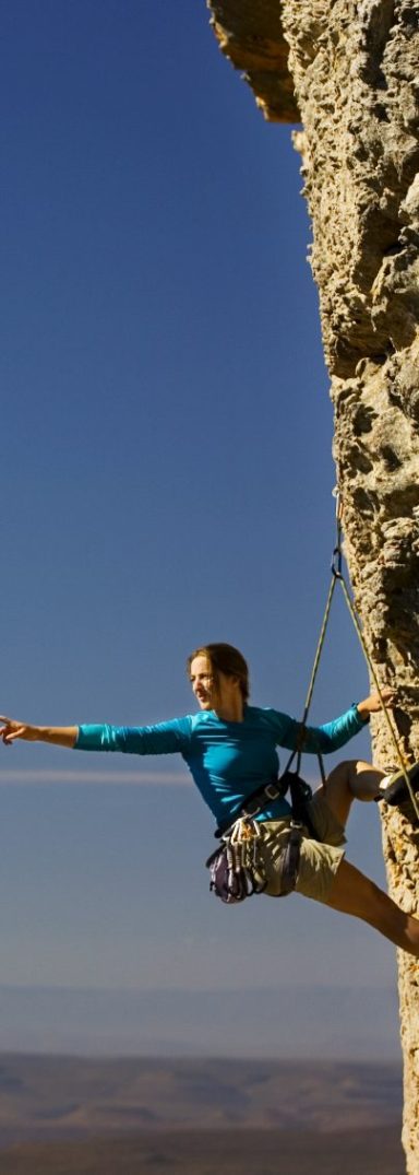 Junge Frau hängt hoch in einer senkrechten Felswand, im Ausfallschritt, den Blick von der Wand weg weit auf den Horizont gerichtet, auf diesen zeigend, gesichert durch Klettergurt und Haken.